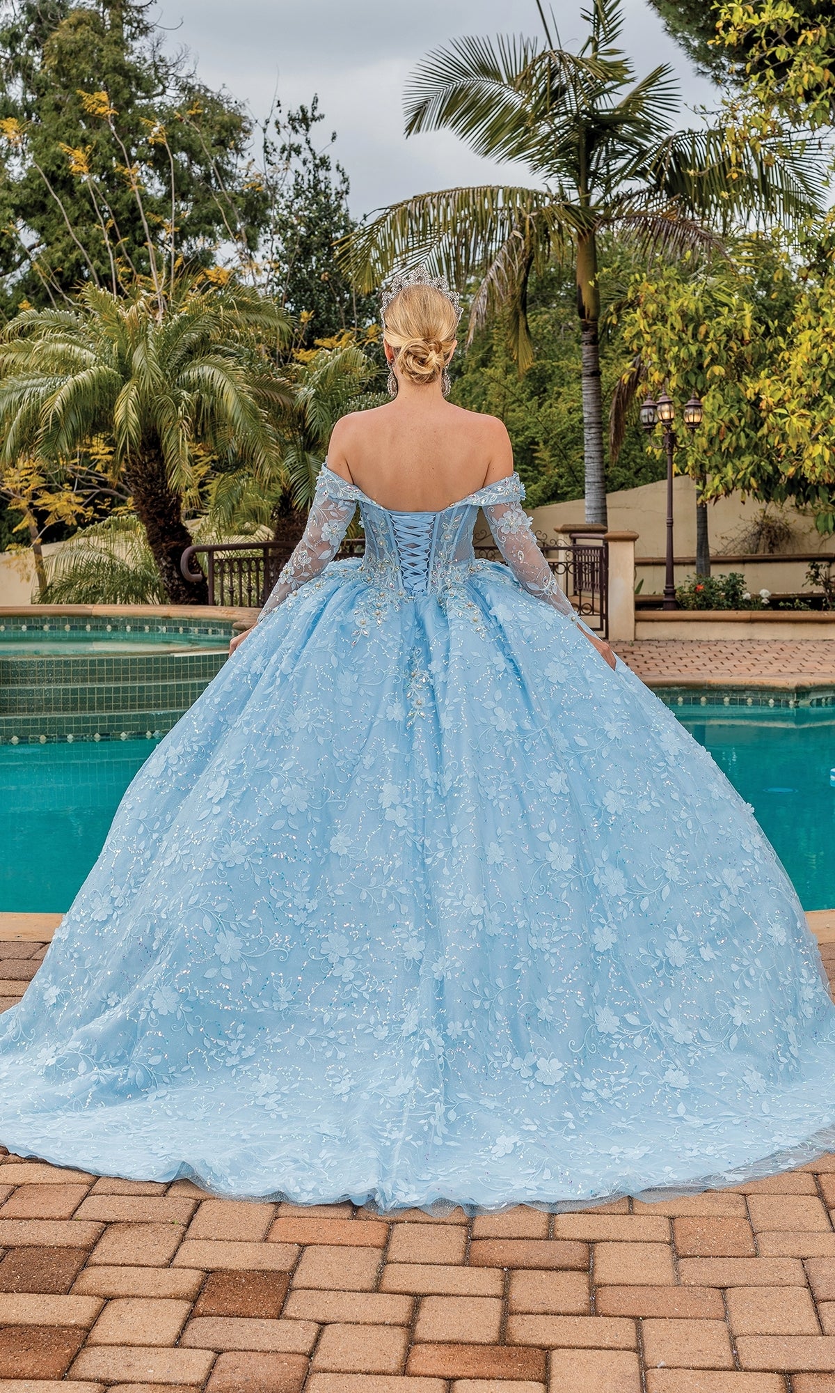 A woman with blonde hair stands by a pool in the Dancing Queen Quinceanera Dress 1890, an elegant light blue off-the-shoulder ball gown. Palm trees sway gently in the background as she faces away from the camera.